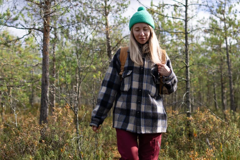 Nainen kävelee metsässä syksyisessä maisemassa, päässään vihreä pipo ja selässään reppu.