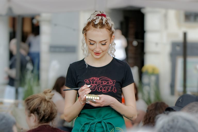 Nuori tarjoilija kirjaa tilausta muistivihkoon ulkoterassilla, yllään musta paita ja vihreä esiliina, taustalla asiakkaita.