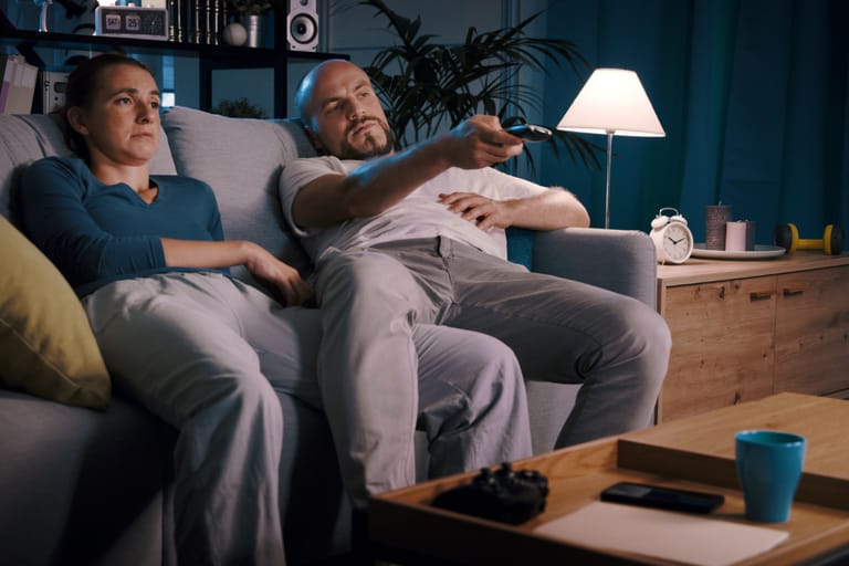 Kaksi aikuista istuu sohvalla olohuoneessa, toinen pitää kädessään kaukosäädintä ja toinen istuu vieressä katsellen eteenpäin