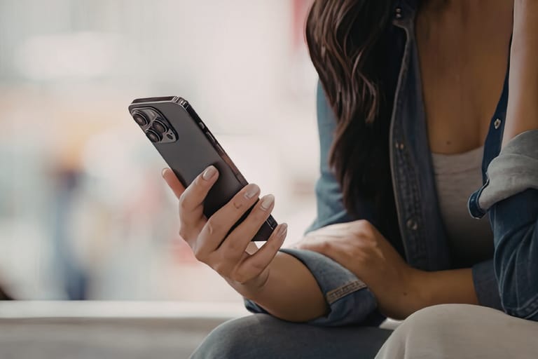 Lähikuva naisesta, joka istuu ja katsoo älypuhelimensa näyttöä pitäen puhelinta toisessa kädessään.