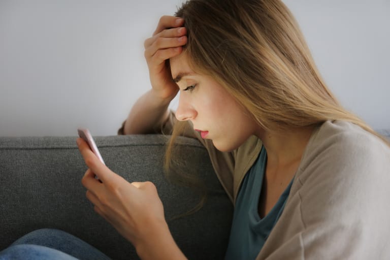 Huolestunut nainen istuu sohvalla ja katsoo puhelintaan käsi otsalla, lähikuva sivuprofiilista vaaleaa seinää vasten.