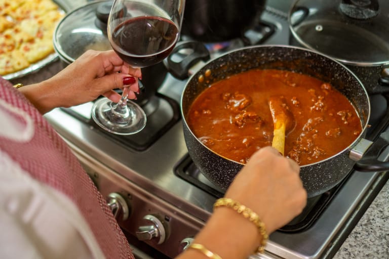 Kattila liedellä, jossa haudutetaan tomaattikastiketta. Vieressä käsi pitelee punaviinilasia.