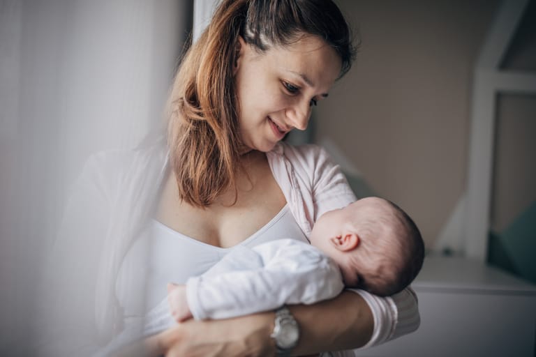 Äiti pitelee vastasyntynyttä vauvaa sylissään ja katsoo tätä lempeästi sisätiloissa rauhallisessa ympäristössä.