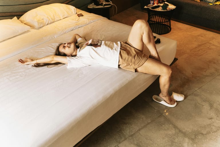 Nainen makaa hotellisängyllä rentoutuneena, jalat sängyn reunalla ja tossut lattialla.