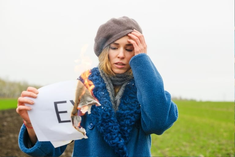 Huolestunut nainen polttaa paperia, jossa lukee “EX”, pellon laidalla.