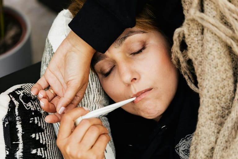 Nainen mittaa kuumetta kuumemittarilla peiton alla kotona