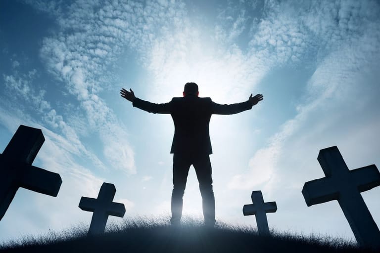 Mies seisoo hautausmaalla ristien keskellä kädet levällään vastavalossa, dramaattinen taivas taustalla