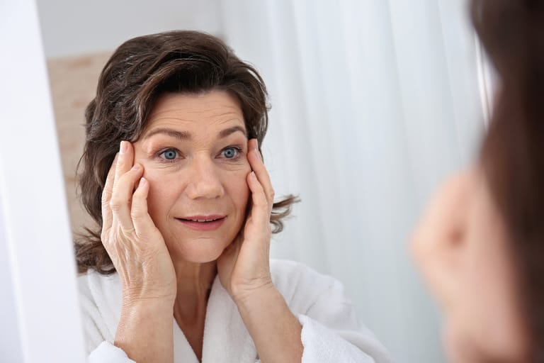 Nainen katsoo peiliin ja koskettaa kasvojaan tarkastellessaan ihoaan.