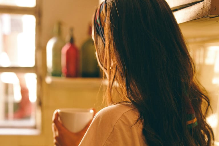 Nainen pitelee kahvikuppia ja katsoo ulos ikkunasta aamunvalossa.