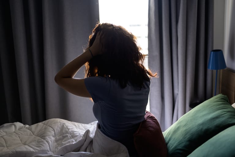Nainen istuu väsyneenä sängyssä aikaisin aamulla ja pitelee päätään.