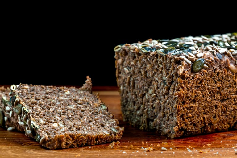 Tumma siemenleipä leikattuna, viipale pöydällä leivän vieressä.