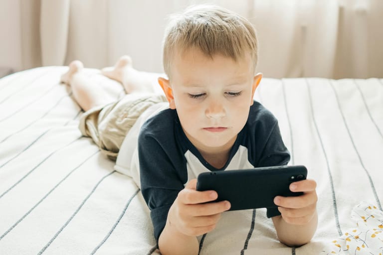Lapsi makaa sängyllä ja katsoo keskittyneenä älypuhelinta.