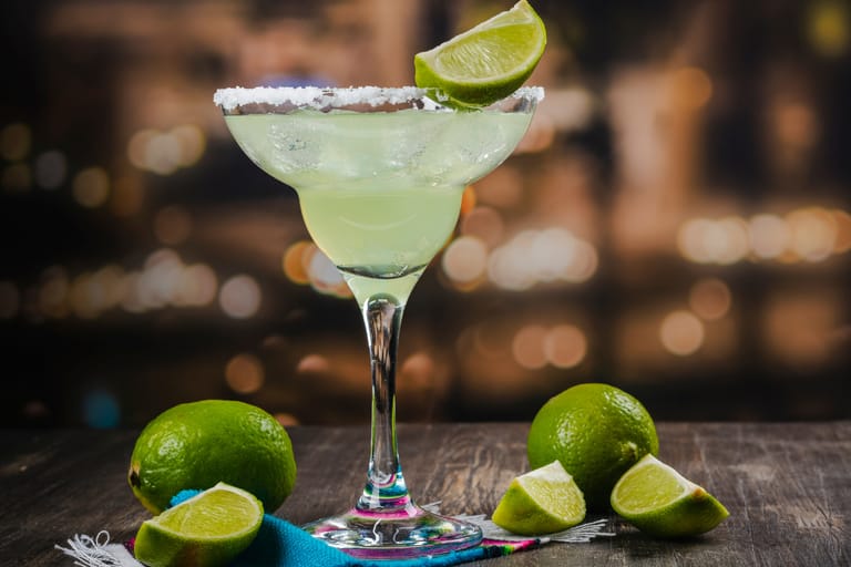 Margarita-cocktail suolareunaisessa lasissa, koristeena limelohko, pöydällä tuoreita limejä taustalla iltavalaistus.