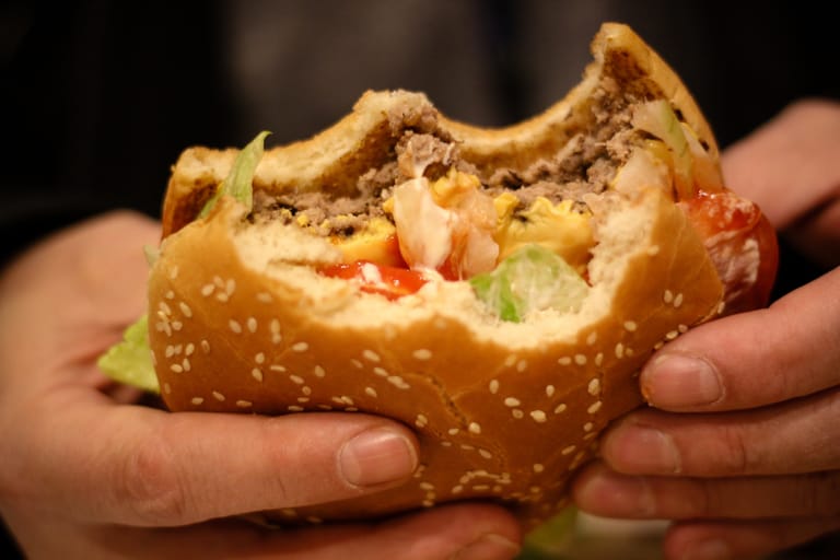 Lähikuva käsissä pidetystä hampurilaisesta, josta on otettu iso puraisu; seesamisämpylän välissä näkyy jauhelihapihvi, juusto, salaatti, tomaatti ja kastike.