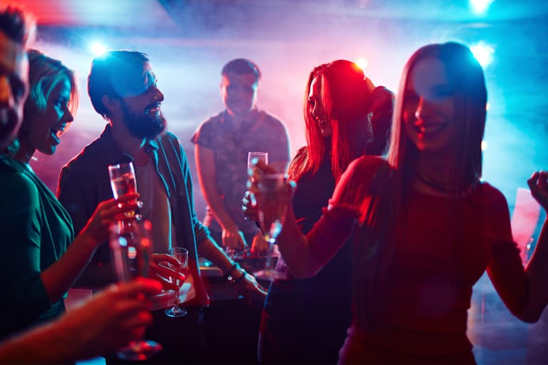 Nuoria aikuisia juhlimassa yökerhossa värikkäiden valojen alla; he tanssivat ja pitävät juomalaseja käsissään DJ:n soittaessa taustalla.