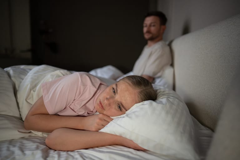 Nainen makaa sängyssä hereillä ja mies istuu taustalla sängyllä.