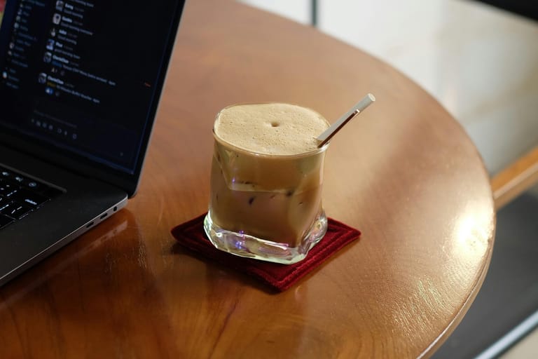 Jääkahvi lasissa pöydällä kannettavan tietokoneen vieressä, juoma on vaahtoisa ja siinä on pilli.