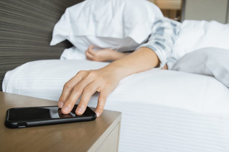 Väsynyt henkilö kurkottaa puhelinta yöpöydältä sängystä käsin.