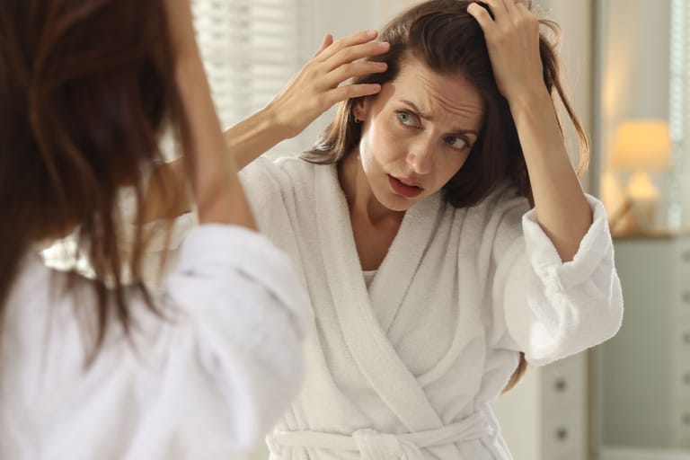 Kylpytakissa oleva nainen katsoo hiuksiaan peilistä ja tutkii hiusrajaansa.