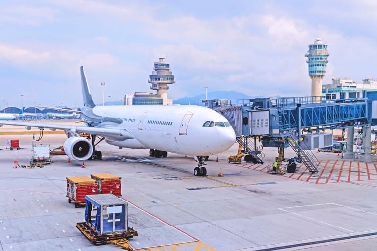 Suuri matkustajalentokone portilla lentokenttäterminaalissa, taustalla lennonjohtotornit ja kuljetuskontteja asfaltilla.