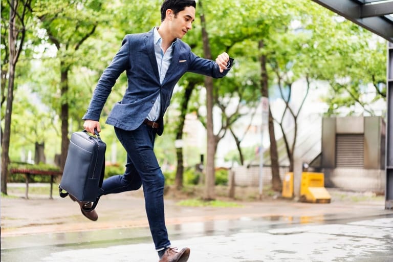 Kiireinen liikemies juoksee sateisella kadulla katsoen kelloa, myöhässä tapaamisesta