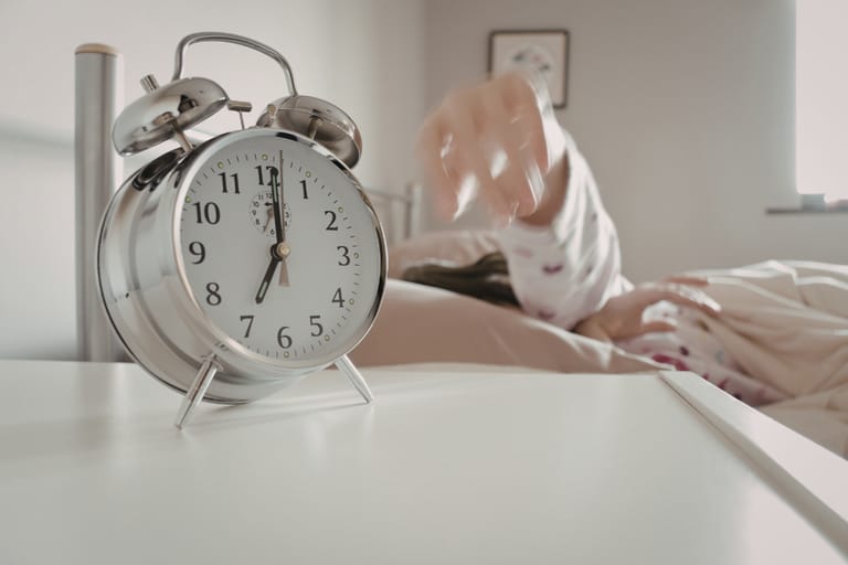 Herätyskello yöpöydällä, käsi kurottaa sitä kohti sängystä.