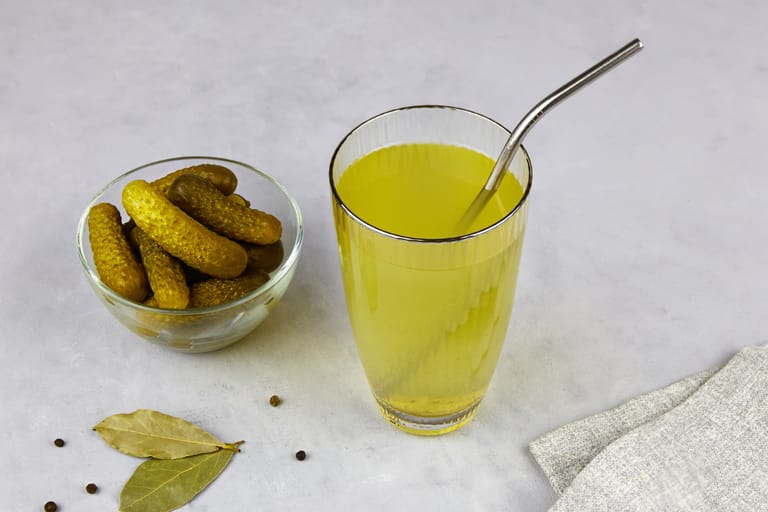 Pöydällä lasi suolakurkkulientä ja vieressä kulho suolakurkkuja.