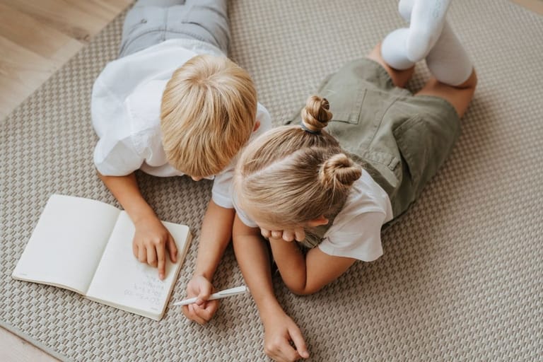 Kaksi vaaleahiuksista lasta makaa lattialla ja tutkii muistikirjaa yhdessä