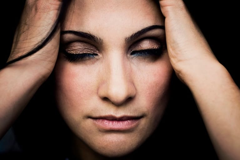 Nuori nainen pitää molempia käsiä ohimoillaan silmät suljettuina mustaa taustaa vasten — ilme kuvastaa uupumusta tai ylikuormitusta