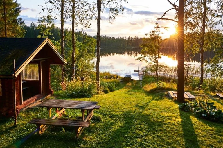 Suomalainen kesämökki järven rannalla iltaruskon valossa, edessä nurmikko ja piknikpöytä, taustalla vene ja koivut.