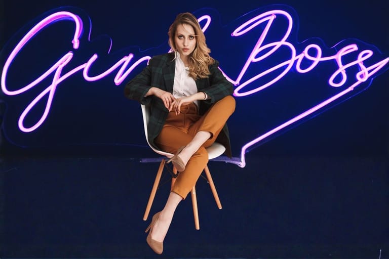 Nuori nainen istuu tuolissa pinkin neon "Girl Boss" -kyltin edessä – tummansinistä taustaa vasten loistava neon-teksti.