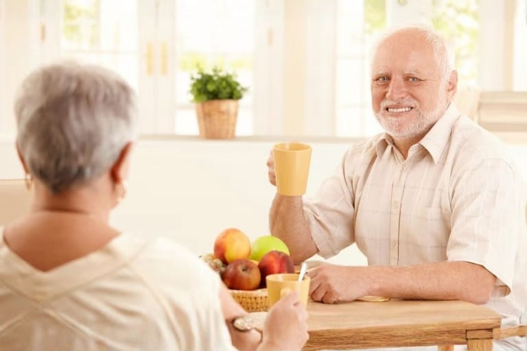 Meemistä tunnettu harmaatukkainen mies, Hide the Pain Harold, istuu keittiön pöydän ääressä pidellen keltaista mukia ja hymyilee vaivaantuneesti naisen suuntaan.