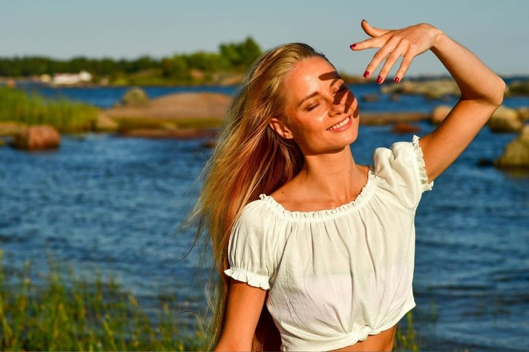 Nainen seisoo silmät kiinni rantakalliolla kesäauringossa