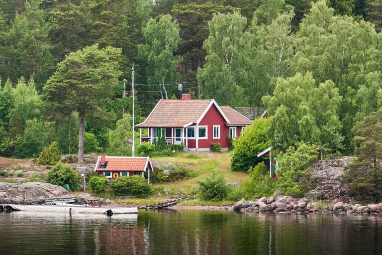 Punainen puinen kesämökki tiilikattoineen sijaitsee vehreässä metsäisessä rinteessä kallioisella järven- tai merenrannalla.