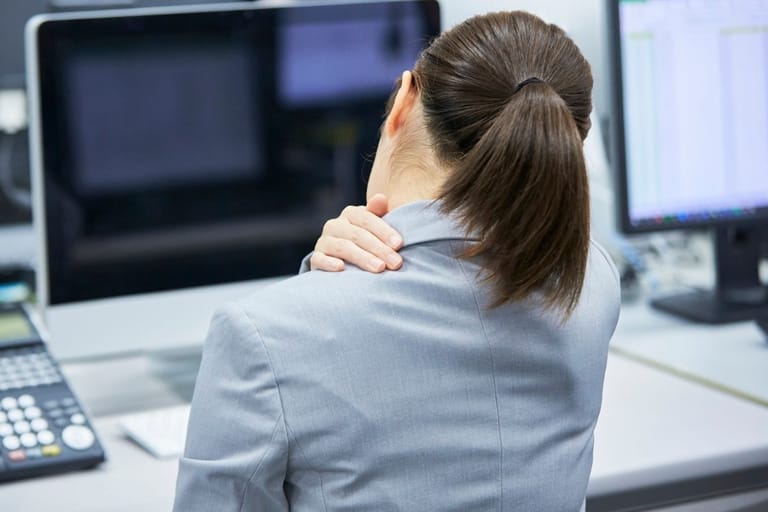 Harmaaseen jakkupukuun pukeutunut nainen istuu työpöydän ääressä toimistossa tietokoneen näyttöjen edessä ja pitelee kipeää niskaansa kädellään.