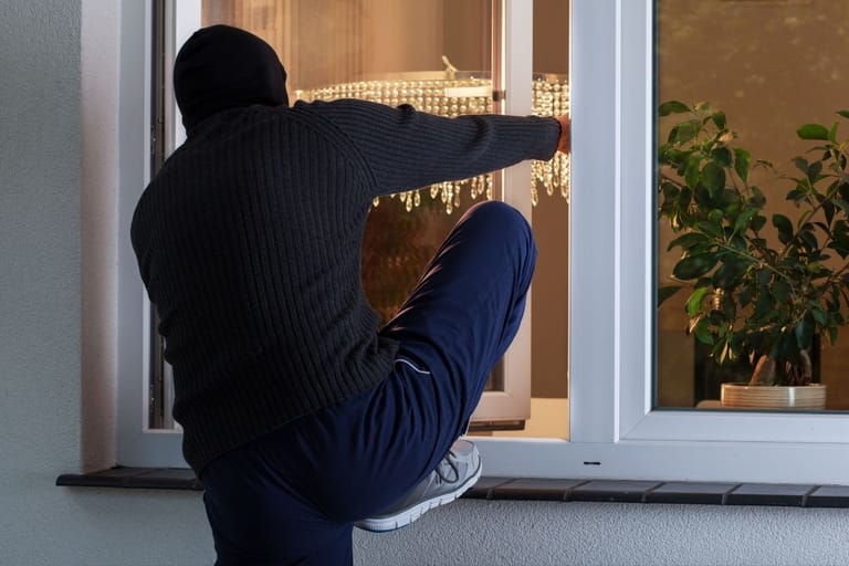 Tummaan huppariin ja mustaan hiihtopipoon pukeutunut murtovaras tunkeutuu sisään avoimesta ikkunasta.