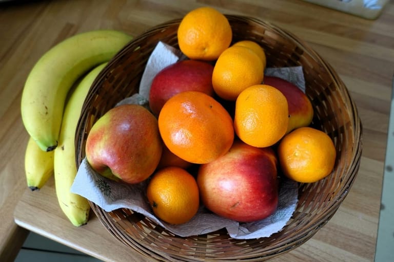 Hedelmäkorissa keittiön pöydällä on omenoita, mandariineja ja mangoja, ja korin vieressä lepää kaksi keltaista banaania.