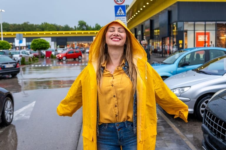 Keltainen sadetakki päällään oleva nainen seisoo hymyillen silmät suljettuina kesäisessä vesisateessa parkkipaikalla autojen keskellä.
