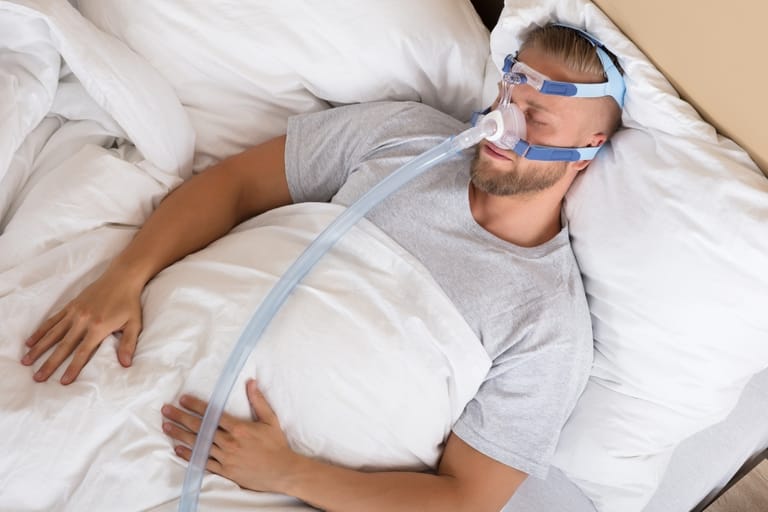 Mies nukkuu selällään CPAP-laitteen maski nenällään.