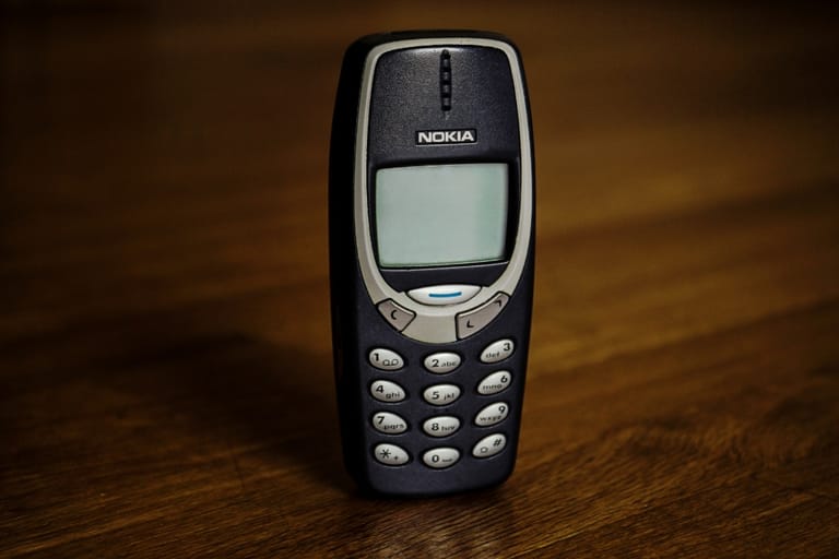 Vanha musta Nokia 3310 -matkapuhelin seisoo puisella pöydällä. Puhelimessa on pieni mustavalkoinen näyttö ja fyysinen numeronäppäimistö.