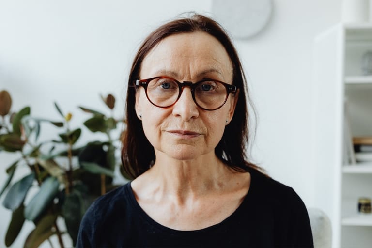 Silmälasipäinen, vakavailmeinen nainen katsoo suoraan kameraan. Taustalla näkyy vaalea huone ja viherkasveja.