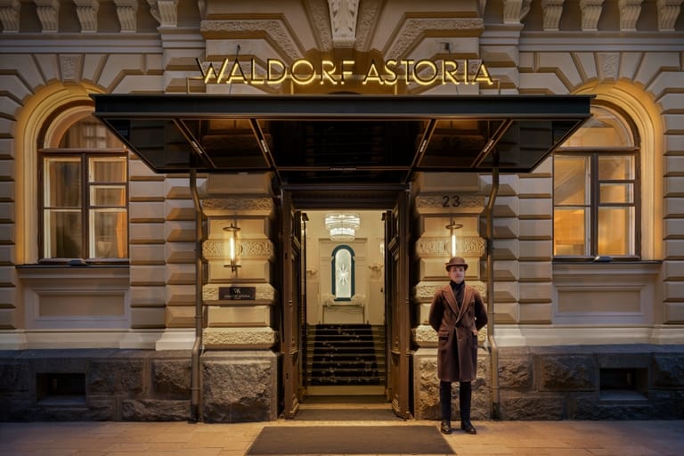 Waldorf Astoria Helsinki -luksushotellin valaistu sisäänkäynti Helsingin Kruununhaassa. Oven edessä seisoo ruskeaan takkiin pukeutunut mies.