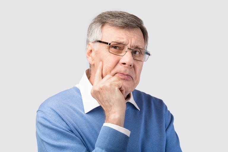 Harmaahiuksinen silmälasipäinen mies katsoo mietteliäänä kameraan sormi poskellaan.