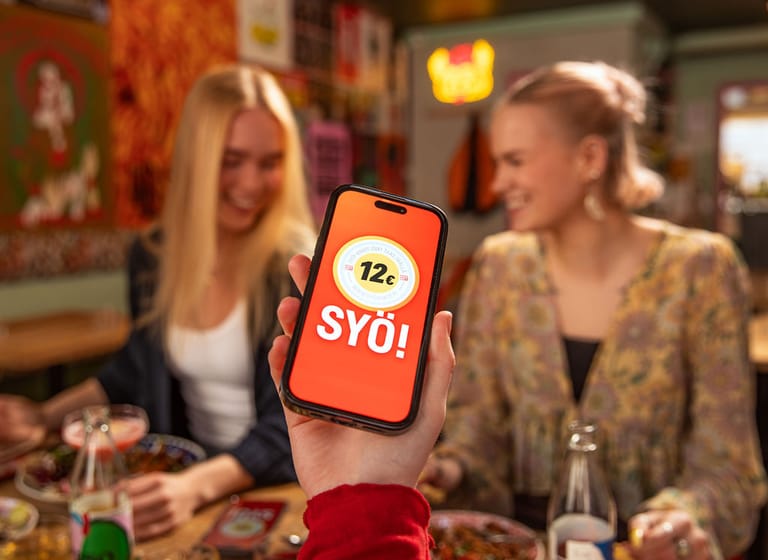 Henkilöitä syömässä ravintolassa. Käsi pitelee puhelinta, jossa näytöllä SYÖ!-viikkojen logo.