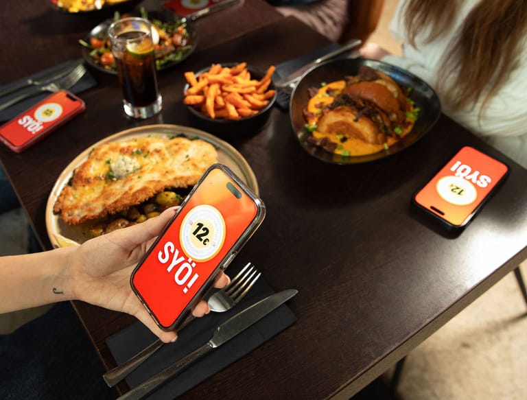 Ravintolapöydässä selataan puhelimista SYÖ!-viikkojen sivua. Taustalla annoksia.