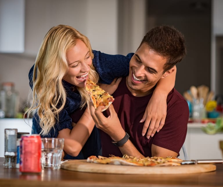 Kuvassa mies ja nainen syövät yhdessä pizzaa romanttisesti. Mies syöttää pizzaa naiselle ja molemmat hymyilevät.