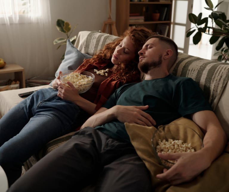 Kuvassa mies ja nainen ovat väsyneinä nukahtaneet yhteisen tv-ohjelman ääreen popcornit sylissä.