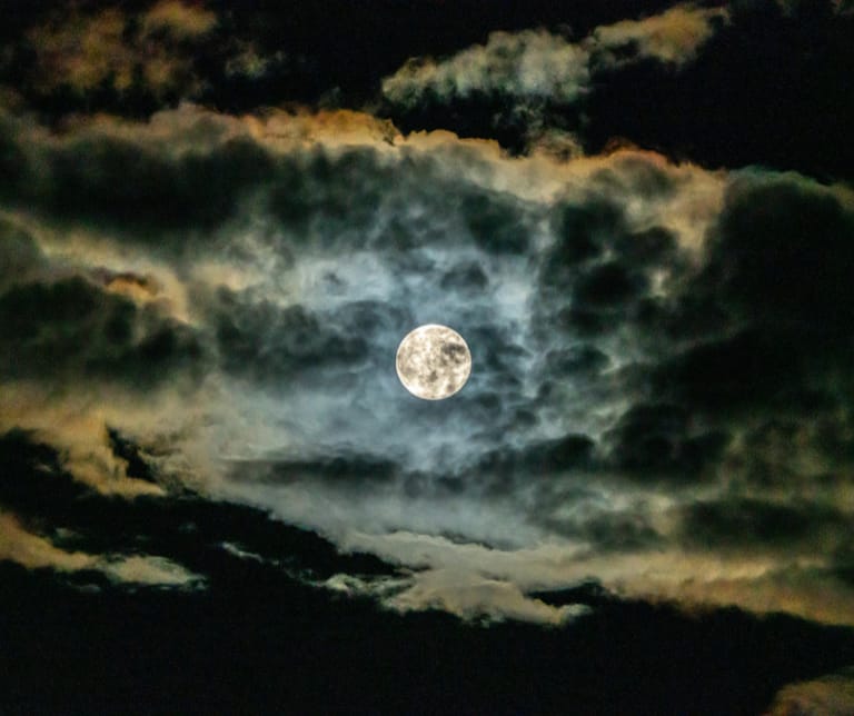 Kuvassa näkyy täysikuu, joka pilkottaa pimeällä taivaalla pilvien takaa.