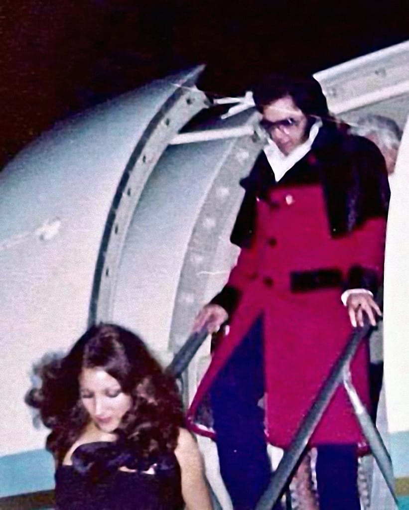 Elvis Presley on nousemassa koneesta aurinkolasit päässään pitkässä punaisessa takissa ja pitelee kiinni lentokoneen nousuportaiden kaiteista. Edellä kulkee Mindi Miller-tyttöystävä. 