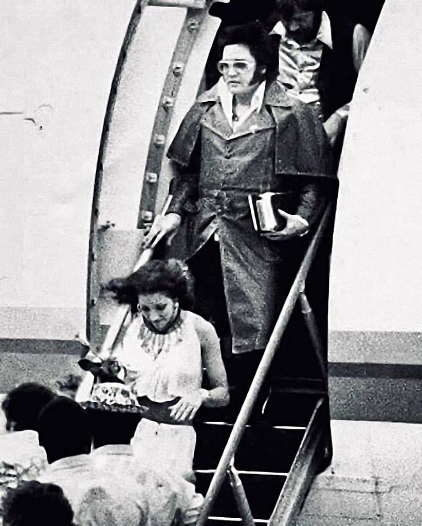 Mindi Miller ja Elvis Presley ovat laskeutumassa koneesta. Mustavalkoinen lehdistökuva, jossa on vahvat 1970-luvun vaikutteet muodissa ja kuvanlaadussa.  